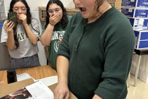 Three students standing around a yearbook pointing at a photo. Their expressions are in surprise. The photo in the yearbook that the students are pointing at is just a group photo of more students. Nothing to be alarmed by, trust me.